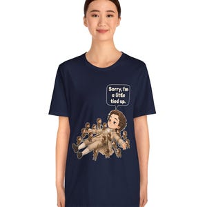 Op de afbeelding: Een marineblauw T-shirt met korte mouwen en een afbeelding van een persoon in 18e-eeuwse kleding die ligt, omringd door vele kleine figuren die ladders gebruiken om hen vast te binden. Een tekstballon bevat de tekst, Sorry, I'm a little tied up.