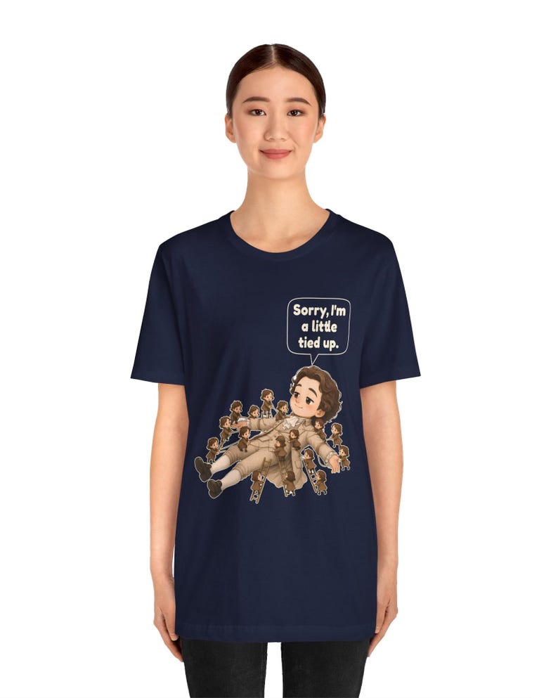 Op de afbeelding: Een marineblauw T-shirt met korte mouwen en een afbeelding van een persoon in 18e-eeuwse kleding die ligt, omringd door vele kleine figuren die ladders gebruiken om hen vast te binden. Een tekstballon bevat de tekst, Sorry, I'm a little tied up.