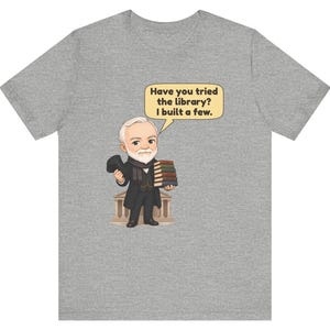 May include: A heather gray t-shirt featuring a cartoon illustration of a bearded man in a suit holding a stack of books and a top hat. A speech bubble above him contains the text: Have you tried the library? I built a few.