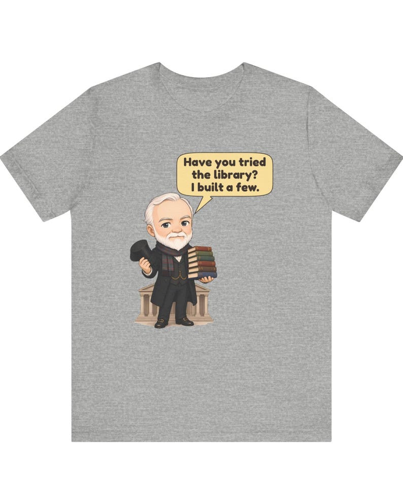 May include: A heather gray t-shirt featuring a cartoon illustration of a bearded man in a suit holding a stack of books and a top hat. A speech bubble above him contains the text: Have you tried the library? I built a few.