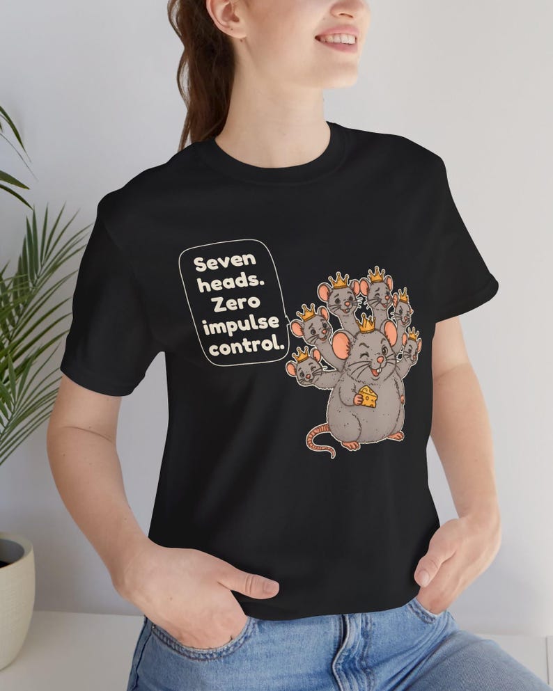 May include: A black t-shirt featuring a graphic of a gray mouse with seven heads, each wearing a small gold crown. The mouse holds a piece of yellow cheese. To the left, a white speech bubble contains the text: Seven heads. Zero impulse control.
