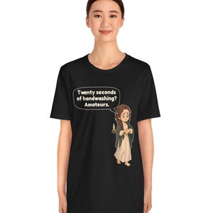Op de afbeelding: Een zwart t-shirt met korte mouwen met een afbeelding van een persoon met bruin haar in een lange jurk die een kaars vasthoudt. Een tekstballon boven het personage bevat de tekst: Twenty seconds of handwashing? Amateurs.
