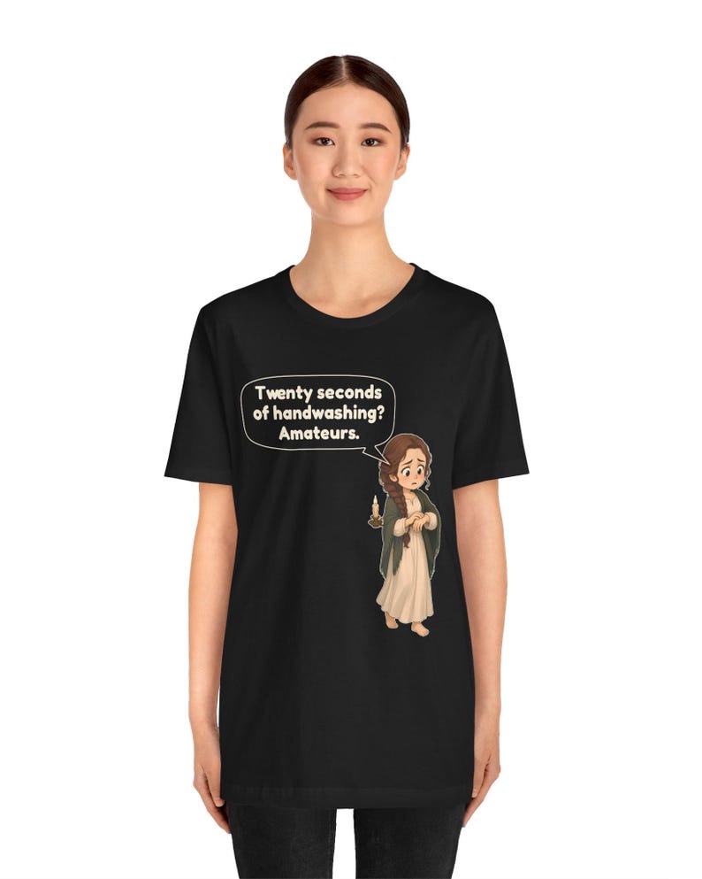 Op de afbeelding: Een zwart t-shirt met korte mouwen met een afbeelding van een persoon met bruin haar in een lange jurk die een kaars vasthoudt. Een tekstballon boven het personage bevat de tekst: Twenty seconds of handwashing? Amateurs.