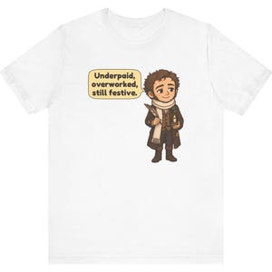 May include: A white short-sleeved t-shirt featuring a cartoon illustration of a person in Victorian-style clothing holding a quill and a candle. A speech bubble next to the character contains the text: Underpaid, overworked, still festive.