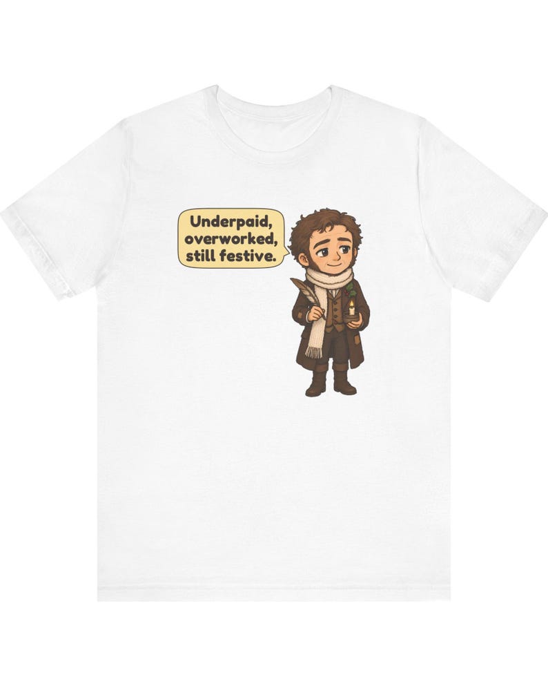 May include: A white short-sleeved t-shirt featuring a cartoon illustration of a person in Victorian-style clothing holding a quill and a candle. A speech bubble next to the character contains the text: Underpaid, overworked, still festive.