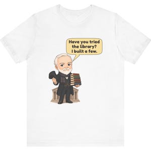 May include: A white t-shirt featuring a cartoon illustration of an older man with a white beard, wearing a black coat and holding a stack of books. A speech bubble above him contains the text: Have you tried the library? I built a few.