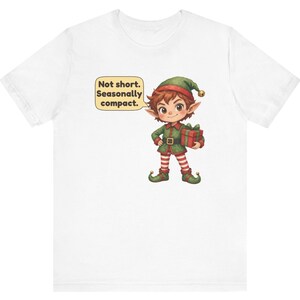 May include: A white short-sleeve t-shirt featuring a graphic of a cartoon elf wearing a green outfit and a hat with a bell. The elf holds a red gift box and has a speech bubble that reads, Not short. Seasonally compact.