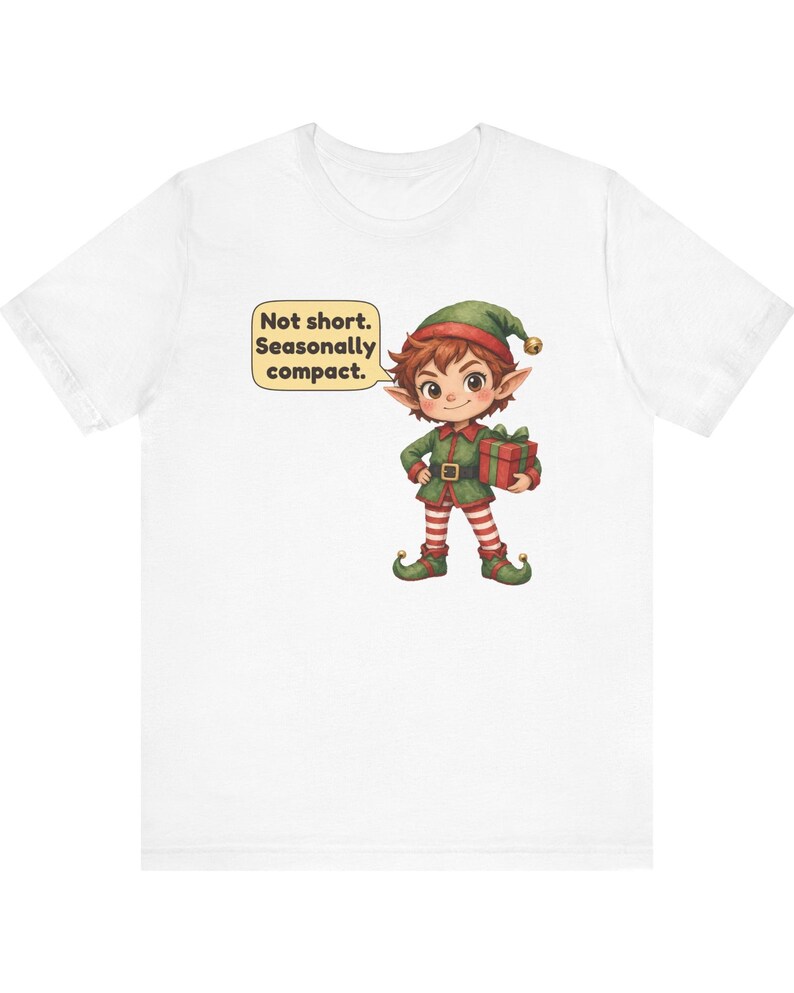 May include: A white short-sleeve t-shirt featuring a graphic of a cartoon elf wearing a green outfit and a hat with a bell. The elf holds a red gift box and has a speech bubble that reads, Not short. Seasonally compact.