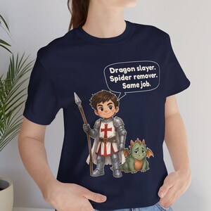 K&ouml;nnte beinhalten: Ein marineblaues T-Shirt mit einer Cartoon-Illustration eines Ritters in silberner R&uuml;stung, der einen Speer h&auml;lt und neben einem kleinen gr&uuml;nen Drachen steht. Eine Sprechblase enth&auml;lt den Text: Dragon slayer. Spider remover. Same job.