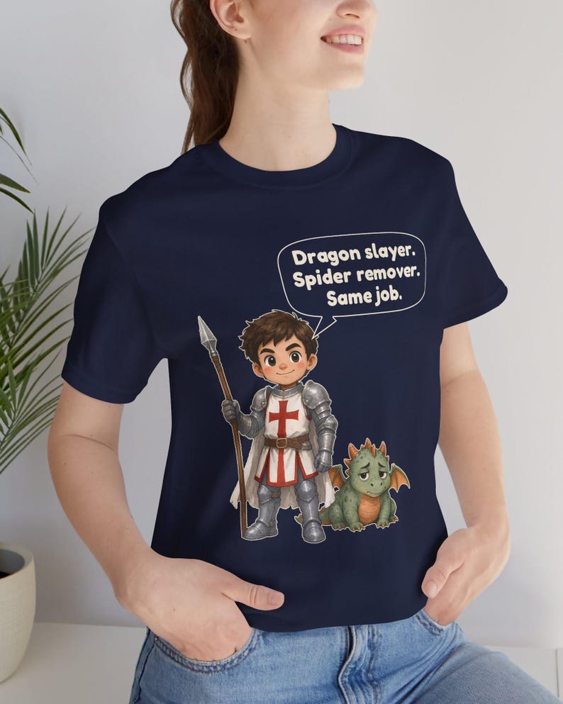 K&ouml;nnte beinhalten: Ein marineblaues T-Shirt mit einer Cartoon-Illustration eines Ritters in silberner R&uuml;stung, der einen Speer h&auml;lt und neben einem kleinen gr&uuml;nen Drachen steht. Eine Sprechblase enth&auml;lt den Text: Dragon slayer. Spider remover. Same job.