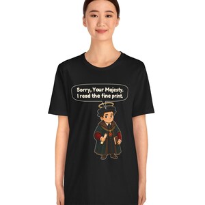 May include: A black t-shirt featuring a graphic of a cartoon character wearing a dark robe, a gold chain, and a halo. The character holds a scroll and a book. Above the character, a speech bubble contains the text: Sorry, Your Majesty. I read the fine print.