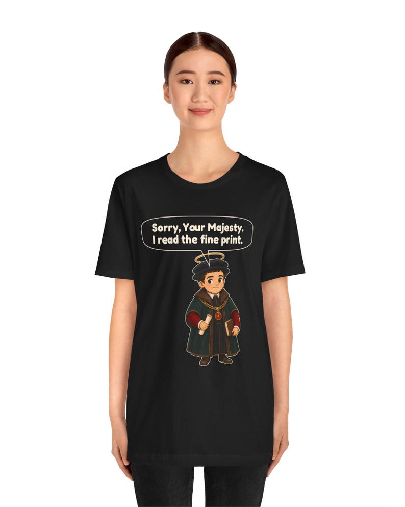 May include: A black t-shirt featuring a graphic of a cartoon character wearing a dark robe, a gold chain, and a halo. The character holds a scroll and a book. Above the character, a speech bubble contains the text: Sorry, Your Majesty. I read the fine print.