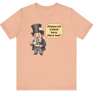 May include: A peach-colored t-shirt featuring a graphic of a character wearing a top hat, a brown vest, and a polka-dot bowtie, holding a teacup. A speech bubble next to the character contains the text: Always six o'clock here. More tea?