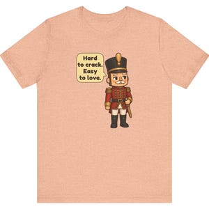 Pu&ograve; includere: Una t-shirt color pesca con l'illustrazione di uno schiaccianoci in uniforme rossa e cappello nero. Un fumetto accanto al personaggio contiene il testo: Hard to crack. Easy to love. Il disegno &egrave; centrato sul petto.