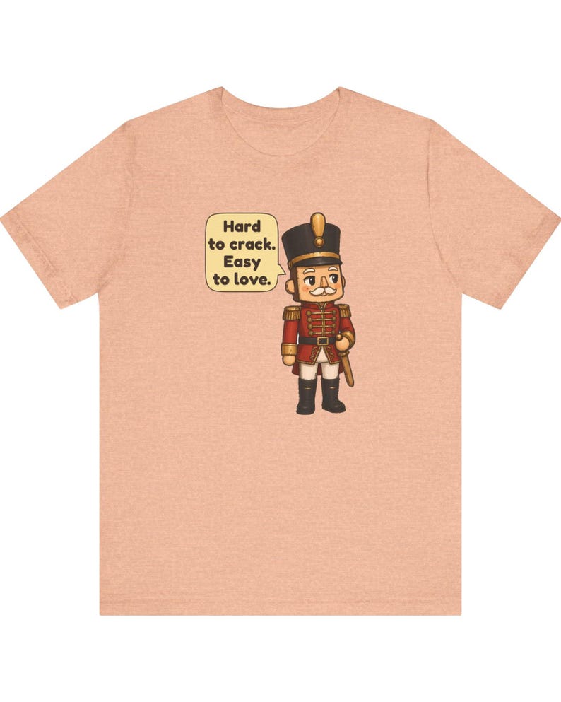 Pu&ograve; includere: Una t-shirt color pesca con l'illustrazione di uno schiaccianoci in uniforme rossa e cappello nero. Un fumetto accanto al personaggio contiene il testo: Hard to crack. Easy to love. Il disegno &egrave; centrato sul petto.