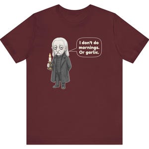 May include: A maroon short-sleeve t-shirt featuring a graphic of a pale, long-haired character in a dark coat holding a lit candle. A speech bubble next to the character contains the text: I don't do mornings. Or garlic.