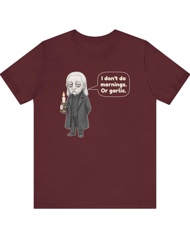 May include: A maroon short-sleeve t-shirt featuring a graphic of a pale, long-haired character in a dark coat holding a lit candle. A speech bubble next to the character contains the text: I don't do mornings. Or garlic.