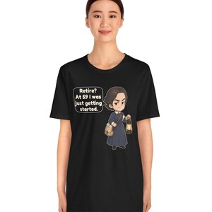 May include: A black t-shirt featuring a cartoon illustration of a person in a dark blue dress holding a lantern and a satchel. A speech bubble next to the character contains the text: Retire? At 59 I was just getting started.