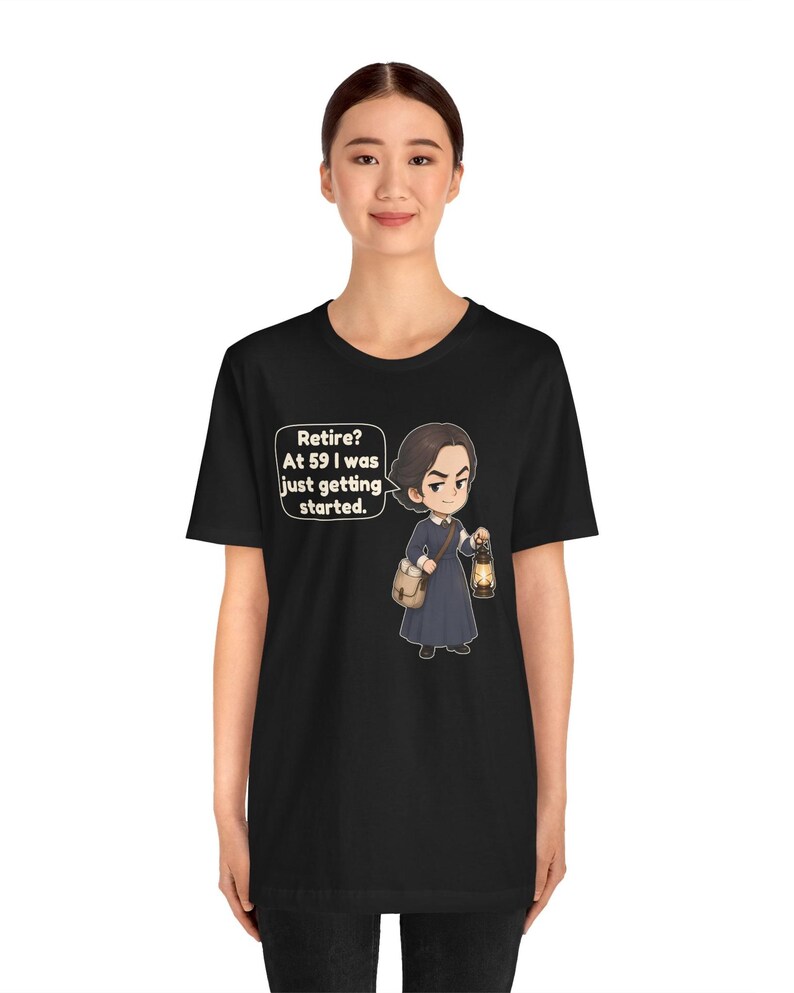 May include: A black t-shirt featuring a cartoon illustration of a person in a dark blue dress holding a lantern and a satchel. A speech bubble next to the character contains the text: Retire? At 59 I was just getting started.