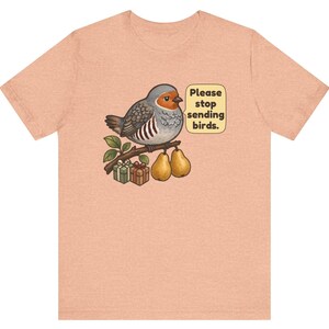 K&ouml;nnte beinhalten: Ein pfirsichfarbenes T-Shirt mit einer Grafik eines grauen und orangefarbenen Vogels, der auf einem Ast mit zwei Birnen und zwei kleinen Geschenkkartons sitzt. Eine Sprechblase neben dem Vogel enth&auml;lt den Text, Please stop sending birds.