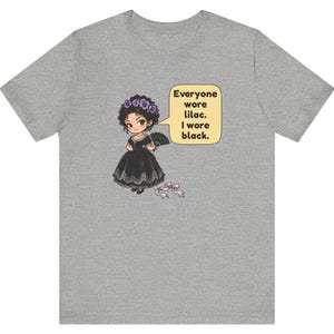 May include: A heather gray t-shirt featuring a cartoon character in a black dress with a lace hem, holding a black fan, wearing a purple flower crown, and a speech bubble that reads: Everyone wore lilac. I wore black. A small purple ribbon lies below.
