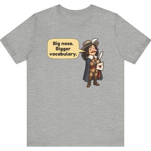 May include: A heather gray t-shirt featuring a cartoon illustration of a historical figure with a large nose, wearing a hat with a feather, a cape, and holding a quill and letter. A speech bubble contains the text: Big nose. Bigger vocabulary.