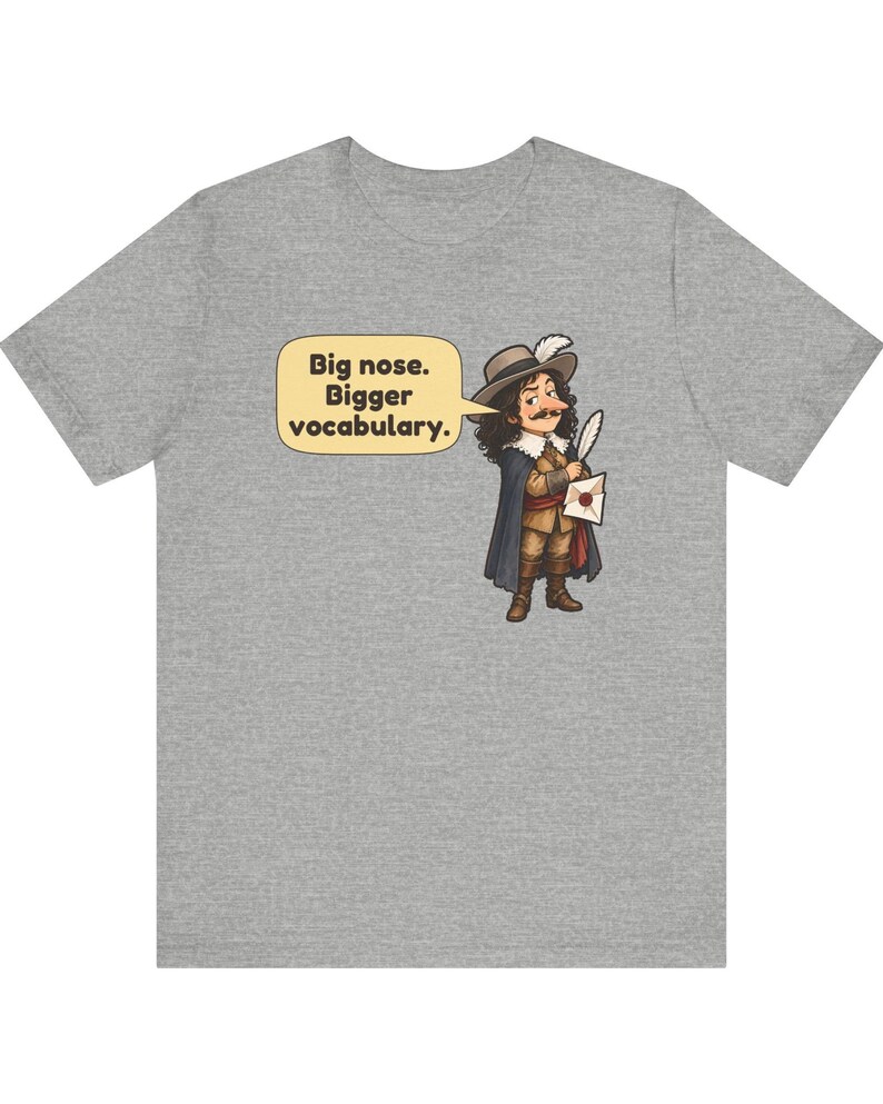 May include: A heather gray t-shirt featuring a cartoon illustration of a historical figure with a large nose, wearing a hat with a feather, a cape, and holding a quill and letter. A speech bubble contains the text: Big nose. Bigger vocabulary.
