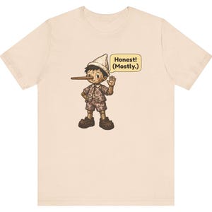 K&ouml;nnte beinhalten: Ein hellbeiges Kurzarm-T-Shirt mit einer grafischen Illustration einer Holzpuppe mit langer Nase, die einen Hut und gemusterte Kleidung tr&auml;gt, sowie einer Sprechblase mit dem Text Honest! (Mostly.)