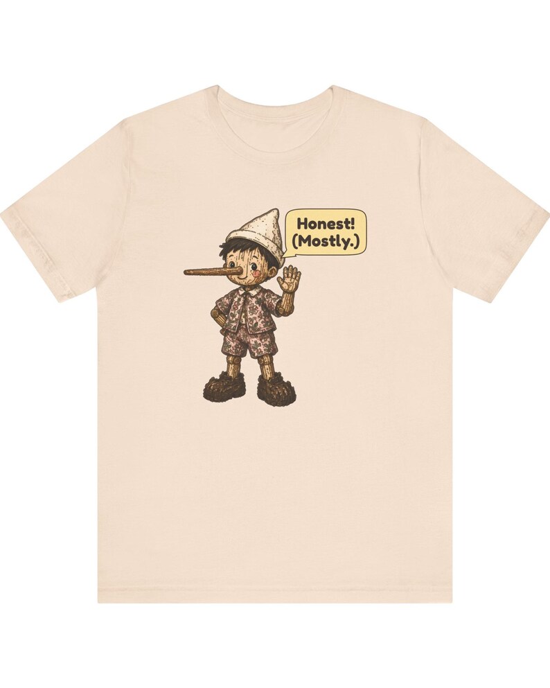 K&ouml;nnte beinhalten: Ein hellbeiges Kurzarm-T-Shirt mit einer grafischen Illustration einer Holzpuppe mit langer Nase, die einen Hut und gemusterte Kleidung tr&auml;gt, sowie einer Sprechblase mit dem Text Honest! (Mostly.)