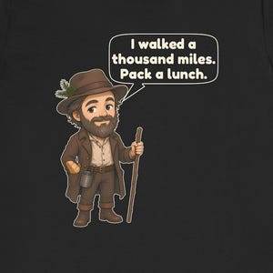 May include: A black t-shirt featuring a cartoon illustration of a bearded hiker wearing a brown hat with a pine sprig, a brown coat, and boots. The hiker holds a wooden walking stick and has a speech bubble that says, I walked a thousand miles. Pack a lunch.
