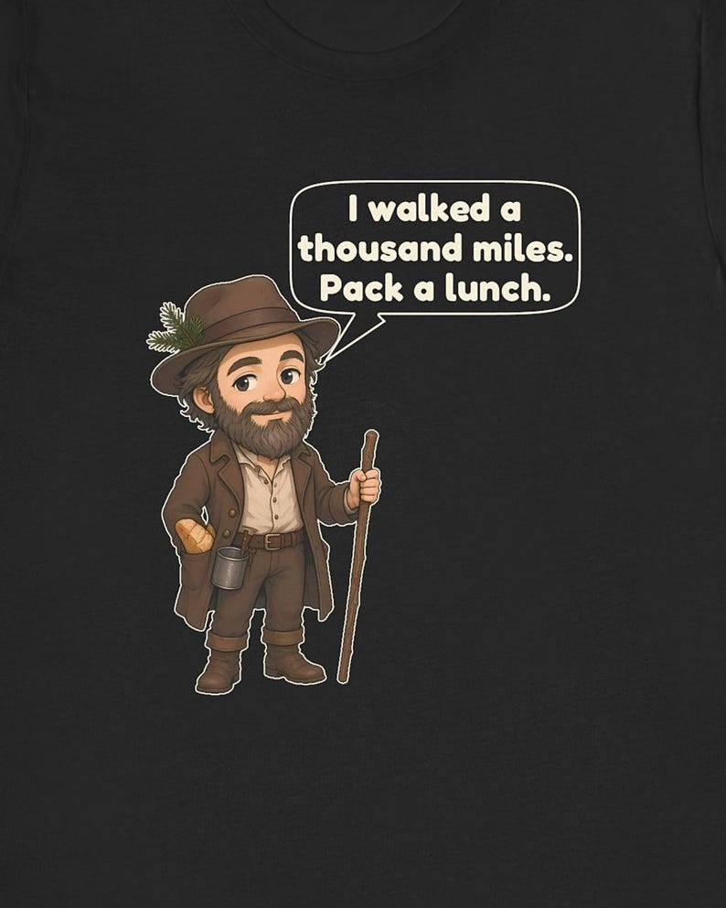 May include: A black t-shirt featuring a cartoon illustration of a bearded hiker wearing a brown hat with a pine sprig, a brown coat, and boots. The hiker holds a wooden walking stick and has a speech bubble that says, I walked a thousand miles. Pack a lunch.