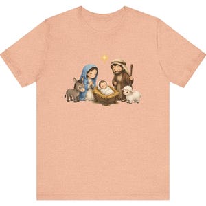 May include: A peach-colored t-shirt featuring a central graphic illustration of a nativity scene. The design depicts a figure in a blue head covering, a figure in a brown robe, a baby in a manger, a donkey, a sheep, and a yellow star.