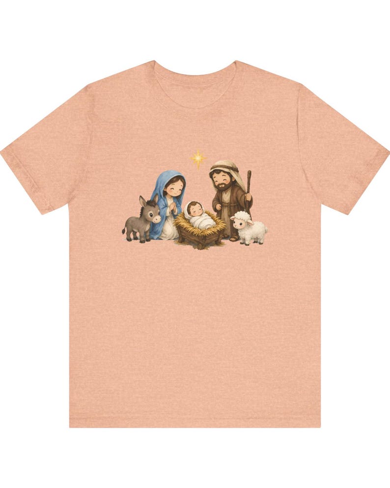 May include: A peach-colored t-shirt featuring a central graphic illustration of a nativity scene. The design depicts a figure in a blue head covering, a figure in a brown robe, a baby in a manger, a donkey, a sheep, and a yellow star.