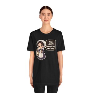 Pode incluir: Uma t-shirt preta de manga curta com um gr&aacute;fico de desenho animado de uma enfermeira com uniforme hist&oacute;rico, segurando uma lanterna e um livro. Um bal&atilde;o de fala cont&eacute;m o texto: Night shift? I brought my own lamp.