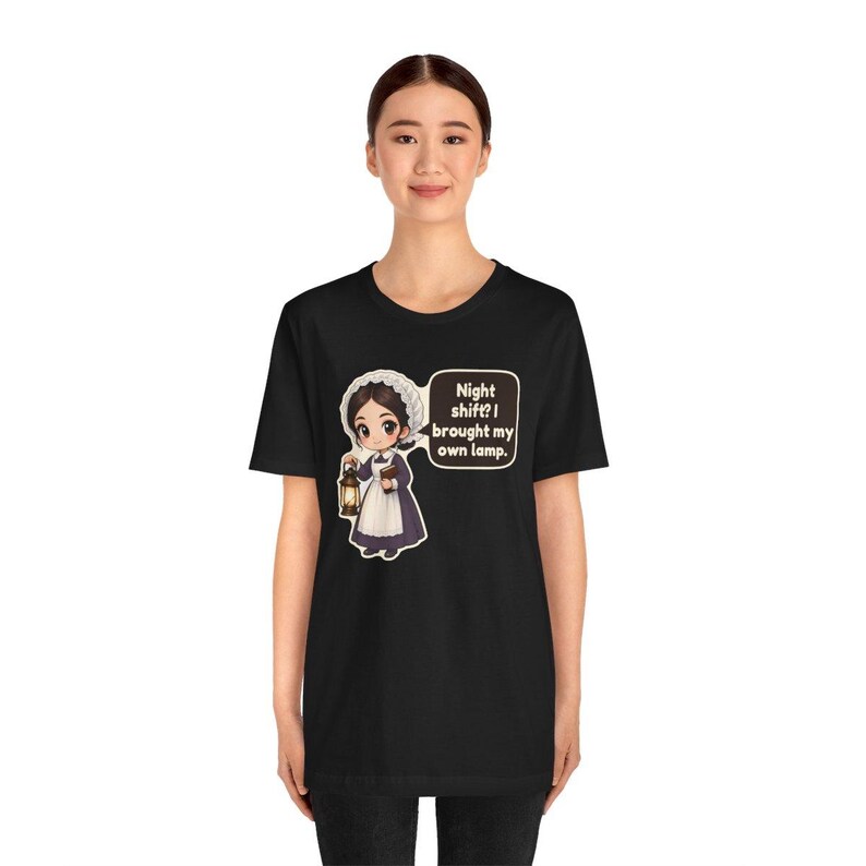 Pode incluir: Uma t-shirt preta de manga curta com um gr&aacute;fico de desenho animado de uma enfermeira com uniforme hist&oacute;rico, segurando uma lanterna e um livro. Um bal&atilde;o de fala cont&eacute;m o texto: Night shift? I brought my own lamp.