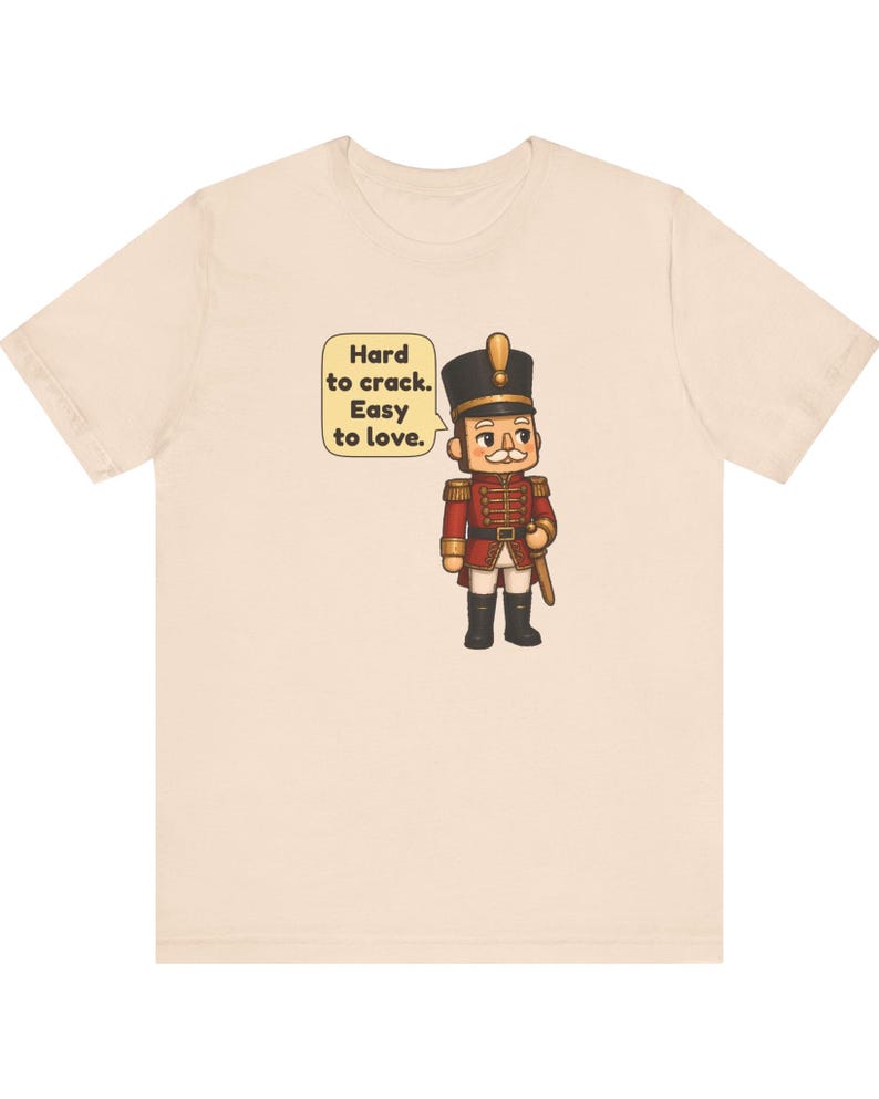 Pu&ograve; includere: Una t-shirt beige chiaro con la grafica di un soldatino schiaccianoci in stile cartoon, con uniforme rossa e cappello nero. Un fumetto accanto al personaggio riporta il testo: Hard to crack. Easy to love. Il disegno &egrave; centrato sul petto.