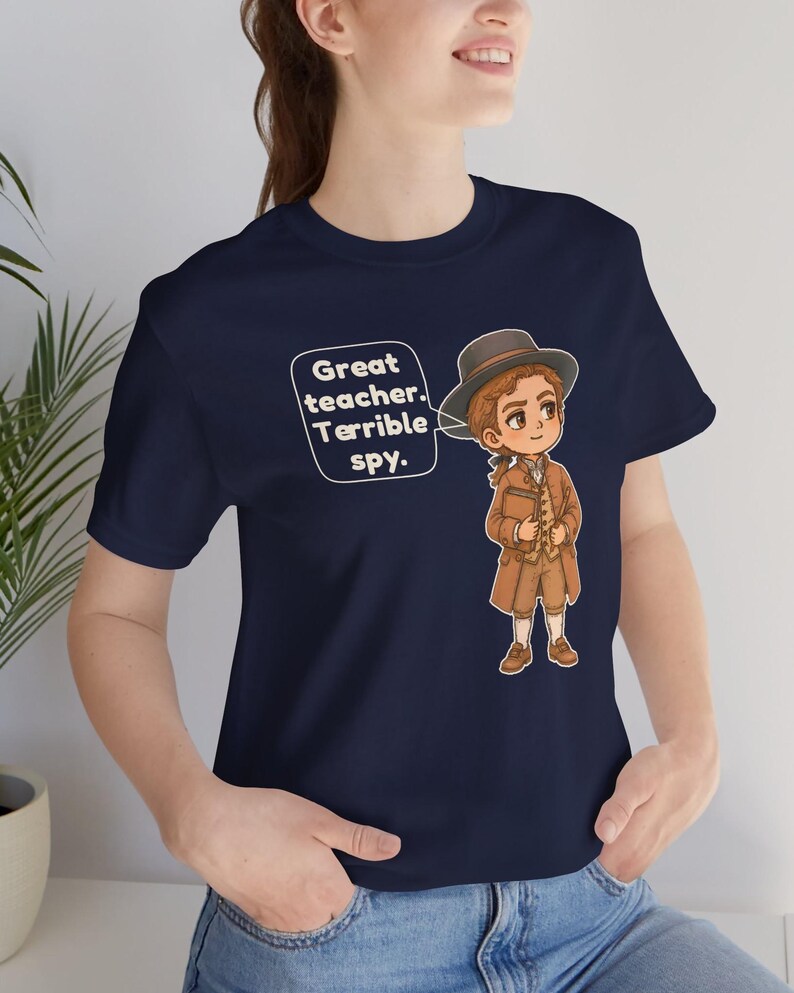May include: A navy blue t-shirt featuring a cartoon illustration of a person in a brown coat and hat holding a book. A speech bubble next to the character contains the text: Great teacher. Terrible spy.