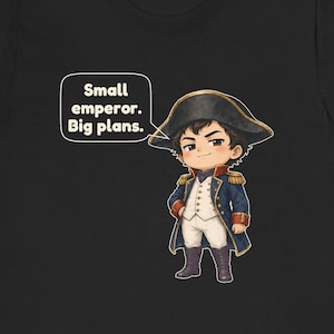 May include: A black t-shirt featuring a cartoon illustration of a small emperor in a blue military coat, white vest, and black bicorne hat. A speech bubble next to the character contains the text: Small emperor. Big plans.