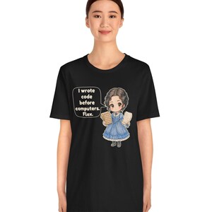 May include: A black t-shirt featuring a graphic of a chibi-style character in a blue Victorian-era dress holding punch cards. A speech bubble next to the character contains the text: I wrote code before computers. Flex.
