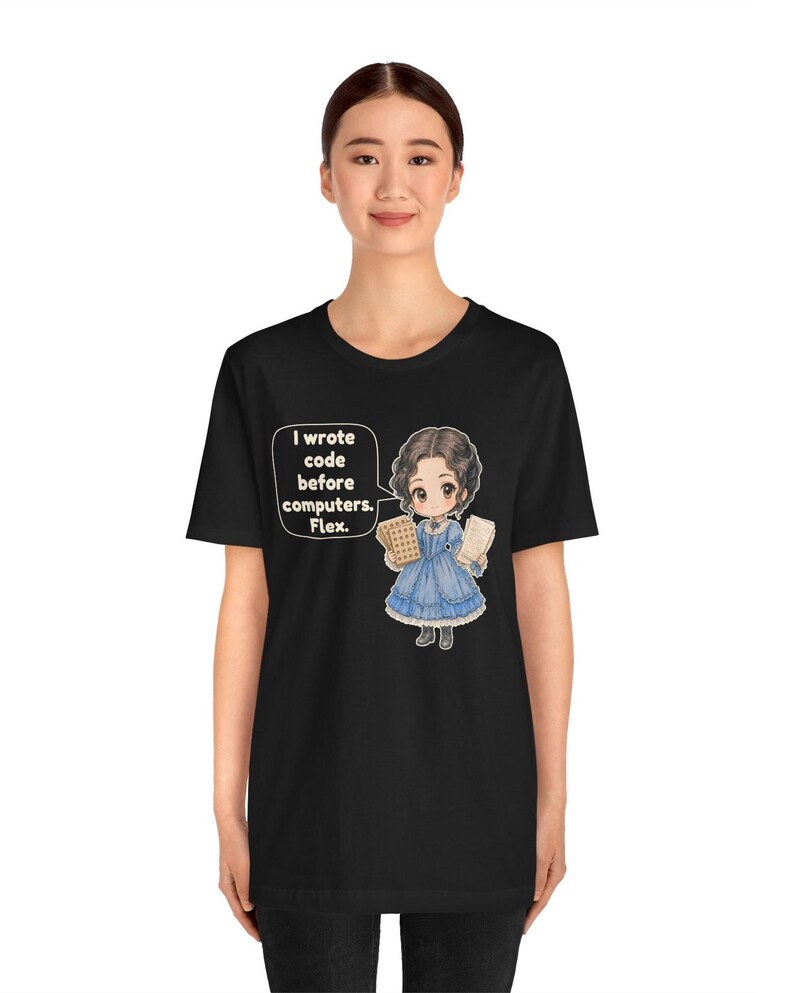 May include: A black t-shirt featuring a graphic of a chibi-style character in a blue Victorian-era dress holding punch cards. A speech bubble next to the character contains the text: I wrote code before computers. Flex.