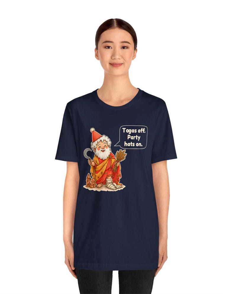 May include: A navy blue short-sleeve t-shirt featuring a graphic of a bearded figure in a red toga and party hat, holding a sickle and wheat. A speech bubble contains the text: Togas off. Party hats on. The figure has a bandaged leg.