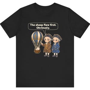 May include: A black t-shirt featuring a graphic of two historical figures in tricorn hats standing next to a hot air balloon with a sheep in the basket. A speech bubble above them contains the text: The sheep flew first. Obviously.