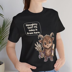 Puede incluir: Una camiseta negra con el gr&aacute;fico de una criatura marr&oacute;n con cuernos, similar a una cabra, que sostiene un manojo de ramas y un pergamino. Un globo de texto sobre la criatura contiene: Naughty list? I'll take it from here.