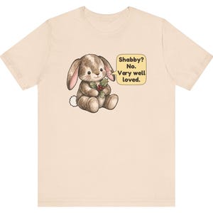 May include: A light beige short-sleeve t-shirt featuring a graphic of a brown stuffed rabbit holding a sprig of holly. A yellow speech bubble next to the rabbit contains the text: Shabby? No. Very well loved.