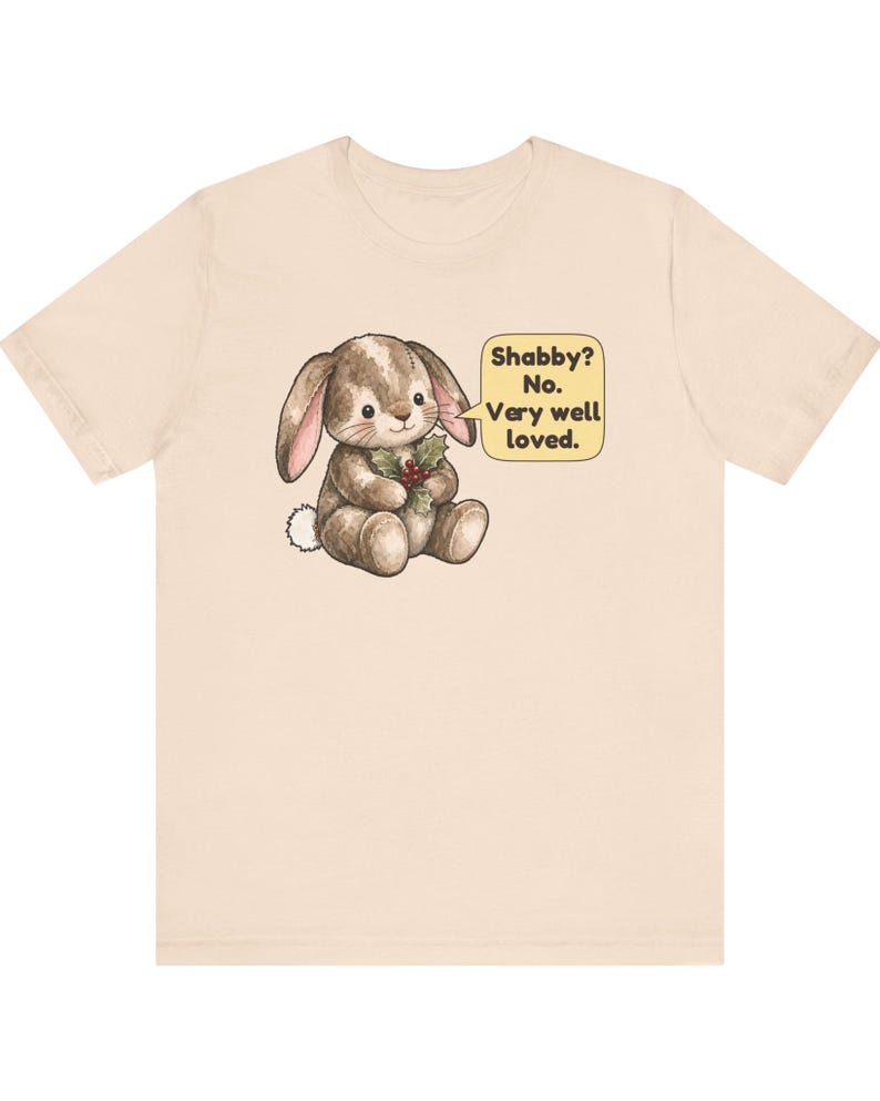 May include: A light beige short-sleeve t-shirt featuring a graphic of a brown stuffed rabbit holding a sprig of holly. A yellow speech bubble next to the rabbit contains the text: Shabby? No. Very well loved.