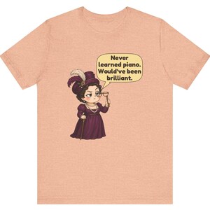 May include: A peach-colored t-shirt featuring a cartoon illustration of a person in a purple Victorian-style dress and hat, holding a monocle. A speech bubble contains the text: Never learned piano. Would've been brilliant.