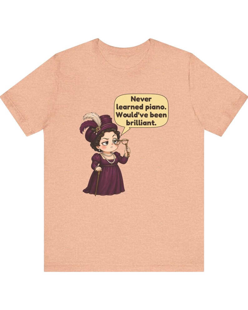 May include: A peach-colored t-shirt featuring a cartoon illustration of a person in a purple Victorian-style dress and hat, holding a monocle. A speech bubble contains the text: Never learned piano. Would've been brilliant.
