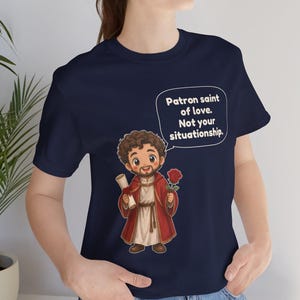 Puede incluir: Una camiseta azul marino con una ilustraci&oacute;n de dibujos animados de una figura con t&uacute;nica roja que sostiene un pergamino y una rosa roja. Un globo de texto contiene: Patron saint of love. Not your situationship.