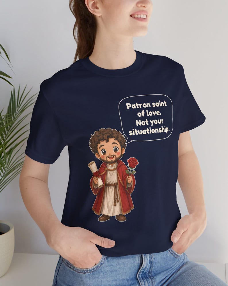 Puede incluir: Una camiseta azul marino con una ilustraci&oacute;n de dibujos animados de una figura con t&uacute;nica roja que sostiene un pergamino y una rosa roja. Un globo de texto contiene: Patron saint of love. Not your situationship.