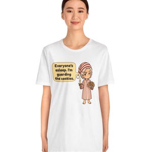 Op de afbeelding: Een wit T-shirt met korte mouwen en een cartoonillustratie van een figuur in een rood-wit gestreepte pyjama die een kaars en een bord koekjes vasthoudt. Een tekstballon bevat de tekst: Everyone's asleep. I'm guarding the cookies.